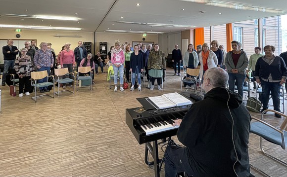Bild: Workshop in einem �bungsraum. Der Chor steht singend kreisf�rmig um den Chorleiter am Piano.
