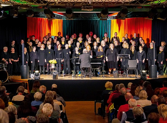 Bild: In der voll mit Besuchern besetzten, bunt beleuchteten Aula der Chor singend auf der B�hne, davor die Besucher begeistert zuh�rend.
