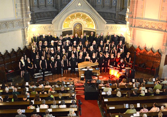 Bild: Von der Kirchenempore von oben nach unten fotografiert sieht man den gro�en und hohen Kirchenraum mit den voll besetzten 
							Kirchenb�nken mit dem Publikum und davor auf der hell beleuchteten B�hne den Chor Get Joy.