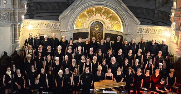 Bild: Von der Kirchenempore herunter fotografiert, sieht man den Chor Get Joy mit ca. 50 S�nger*innen in schwarzer Kleidung 
							singend auf der B�hne vor dem beleuchteten Altar und davor den Chorleiter und den Schlagzeuger.